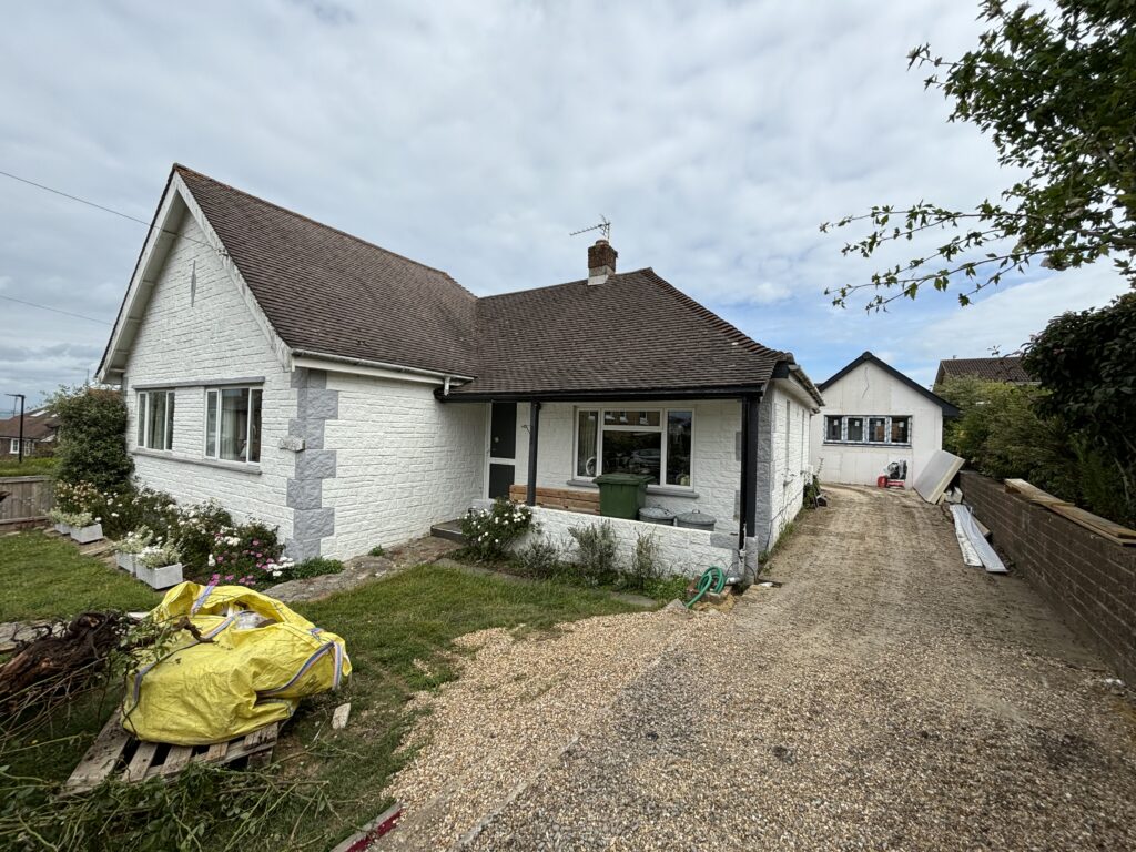 Exterior view of a white bungalow with a gravel driveway, garden area and renovation debris.