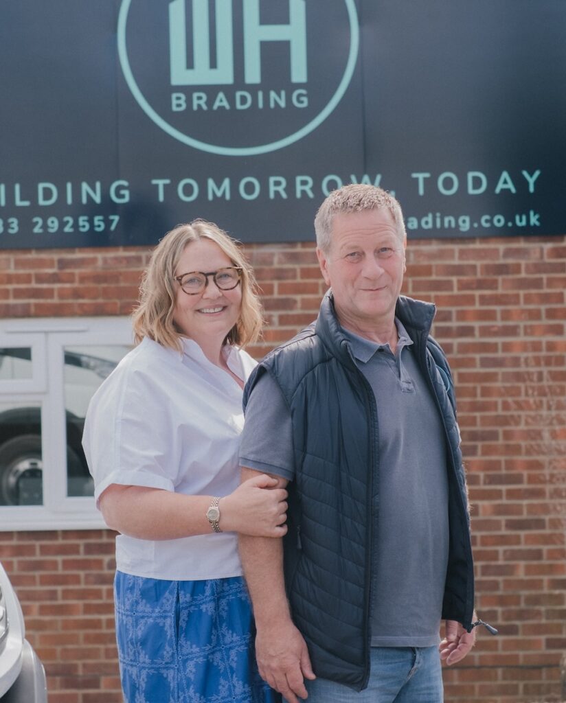 A smiling woman and man standing together in front of a brick building. Behind them is a large black sign featuring a logo and the text "WH BRADING" and "BUILDING TOMORROW, TODAY".